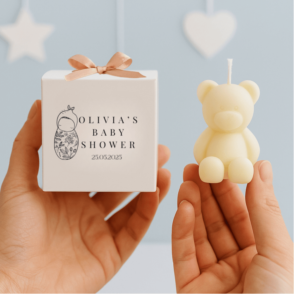 Small bear-shaped candle held by a hand with a decorative engraved box 'Olivia's Baby Shower' in the background.