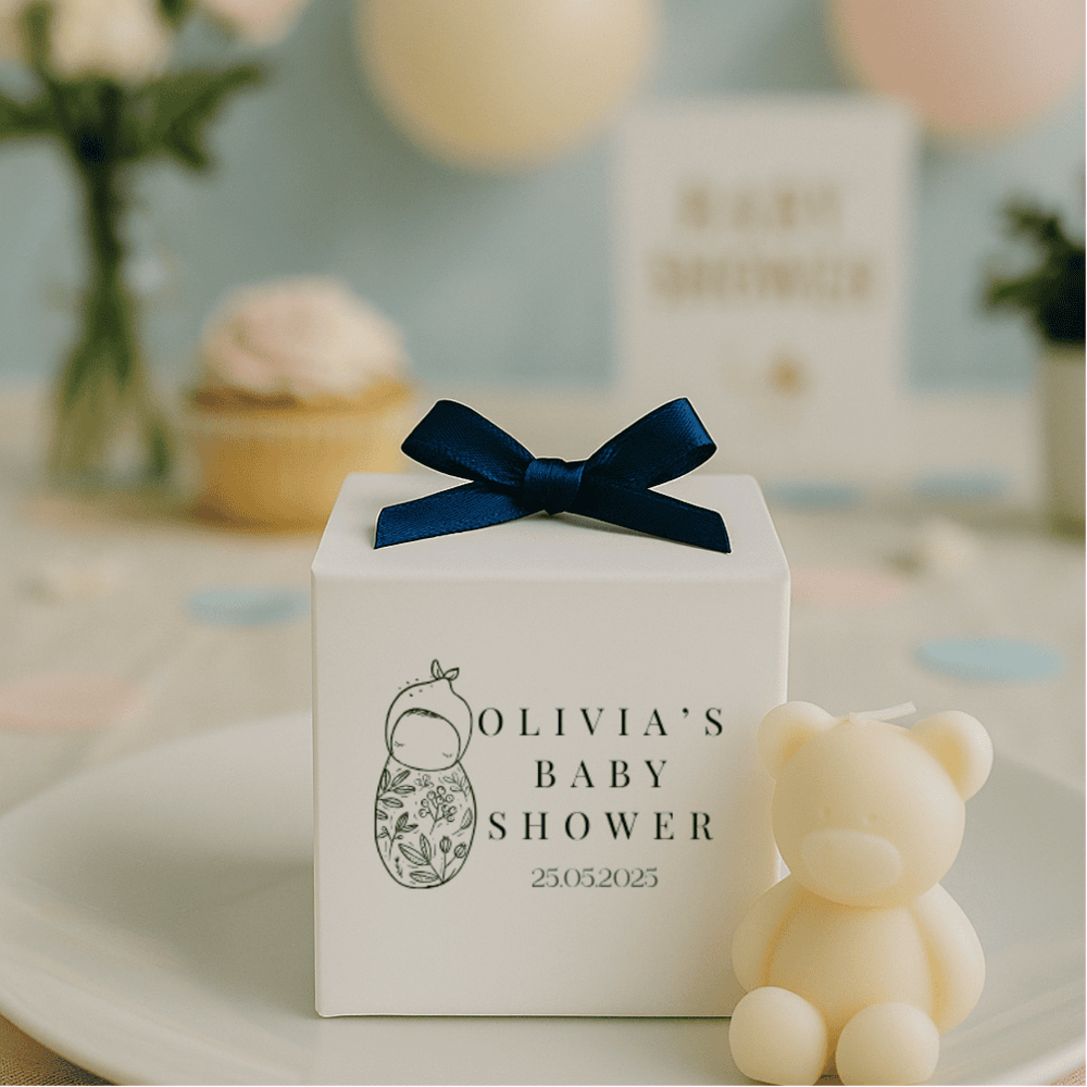White gift box with navy bow and 'Olivia's Baby Shower' text, next to a teddy bear on a decorative table.