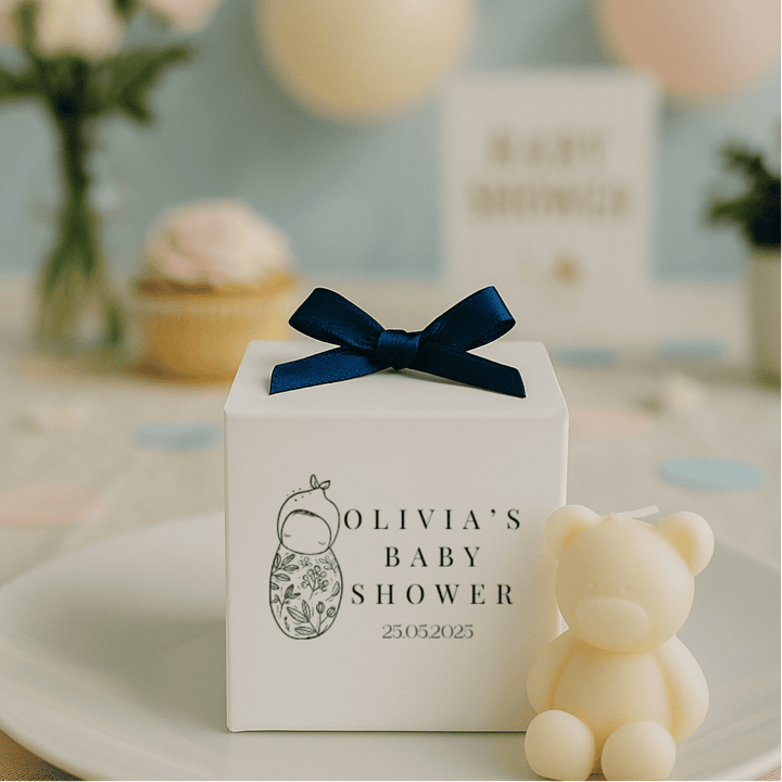 White gift box with navy bow and 'Olivia's Baby Shower' text, next to a teddy bear on a decorative table.