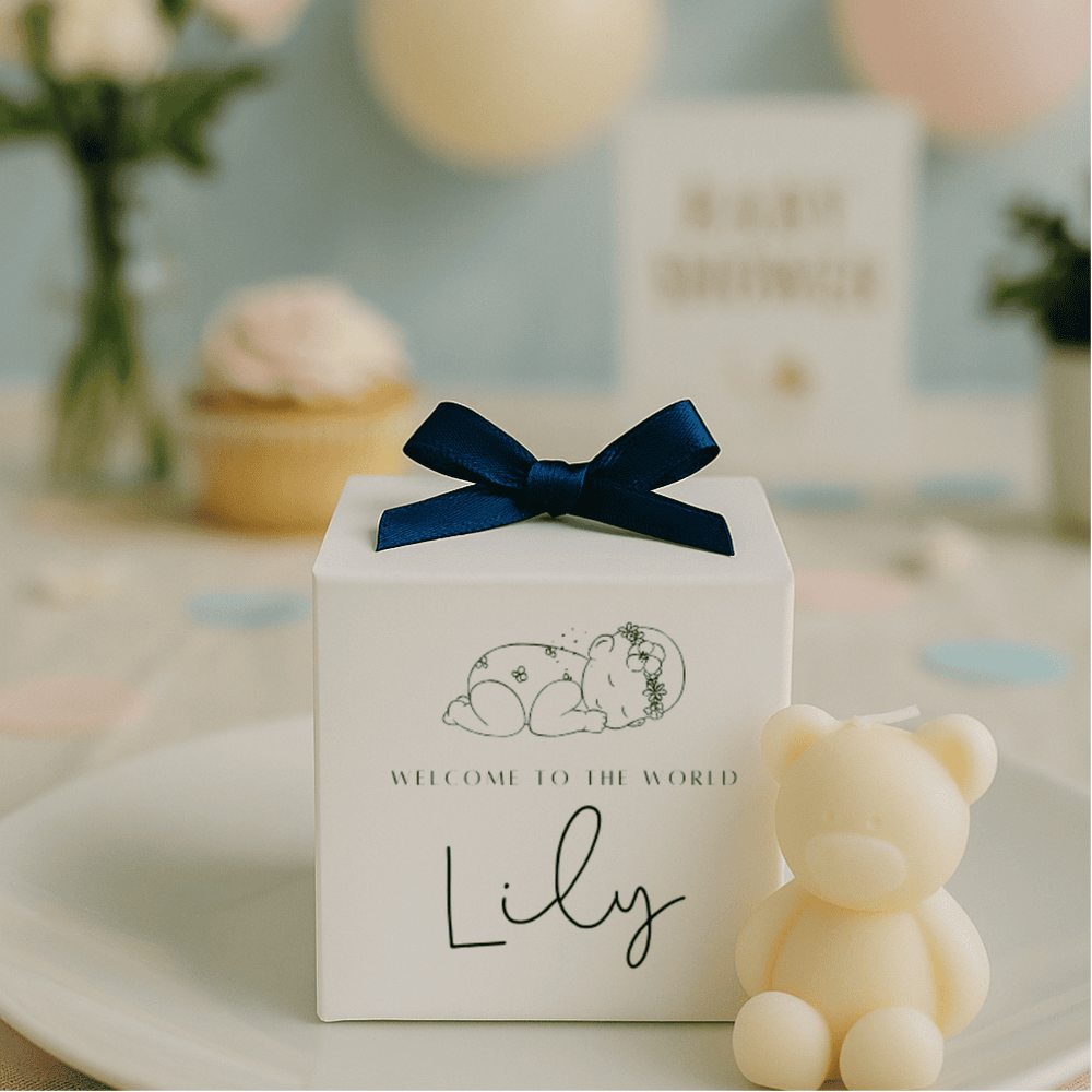 White gift box with navy bow and 'Welcome to the World Lily' text, accompanied by a small teddy bear on a blurred background.