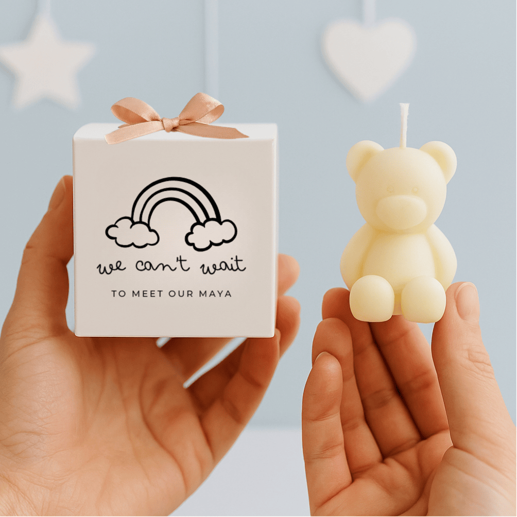 Small white box with 'we can't wait to meet our Maya' text and a bear-shaped candle held by hands against a light blue background with star and heart shapes.